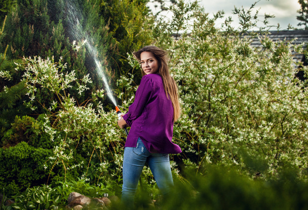 Beautiful joyful young girl in violet casual shirt poses in a summer garden with water hose. - Foto, Imagen