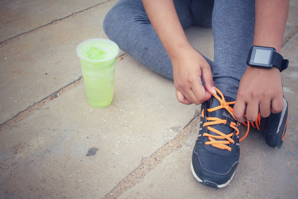Manos de mujer atando cordones de zapatos y smartwatch con batido de verduras verdes
. - Foto, imagen