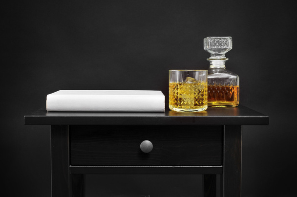 a book, a bottle and a glass with liquor on a table, over a blac - Foto, afbeelding