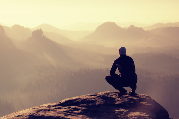 Young man in black sportswear is sitting on cliff edge and looking to misty valley bellow - Photo, Image