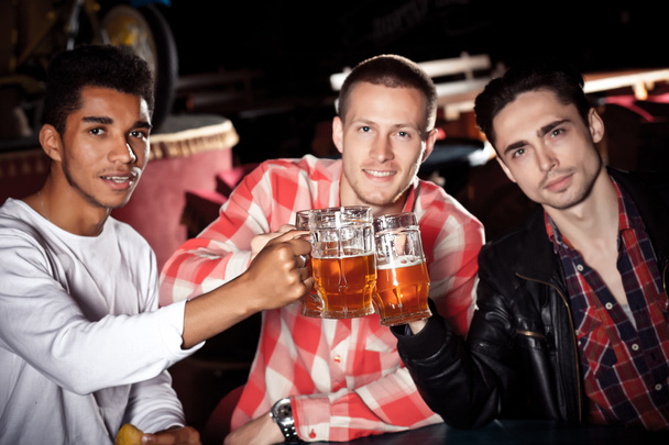 Happy friends clinking with beer mugs in pub, smiling. - Fotó, kép