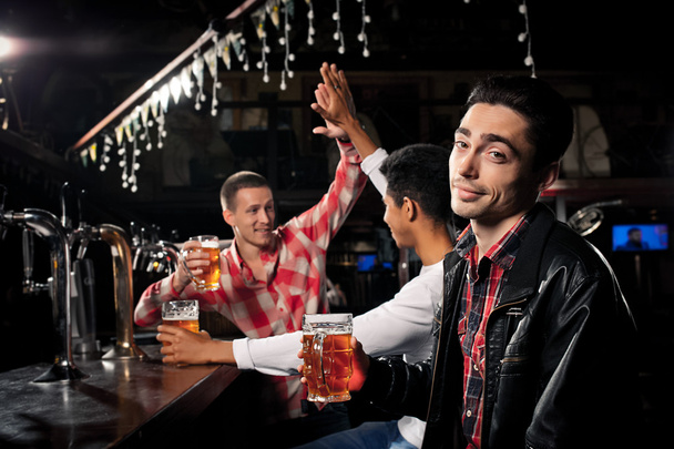 Happy friends clinking with beer mugs in pub, smiling. - Foto, Imagen