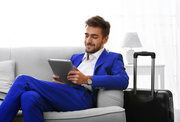 Business man with suitcase and tablet sitting on sofa at home - Фото, изображение