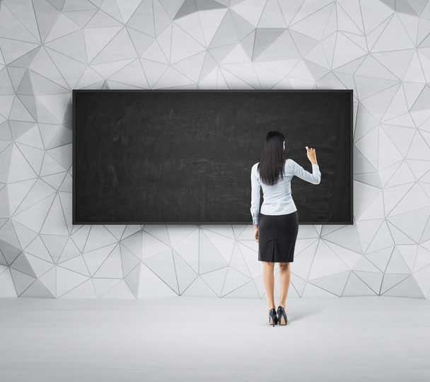 Rear view of full-length brunette woman who is writing something on the empty black board. - Foto, afbeelding