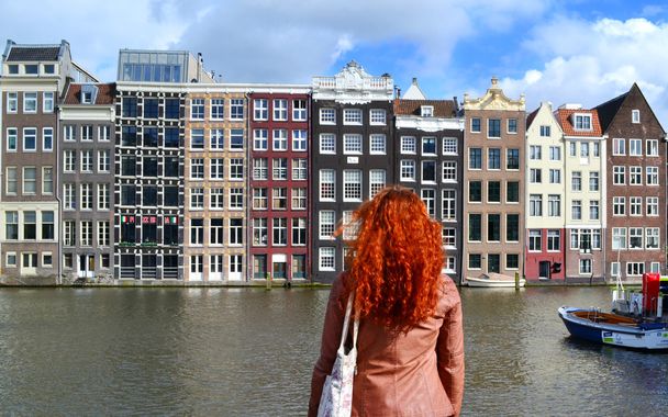 Kamera ile onu geri duran ve kanal ve tekneler yakınındaki Amsterdam şehrin geleneksel yapılar bakarak deri ceketli Kızıl saçlı kadın - Fotoğraf, Görsel