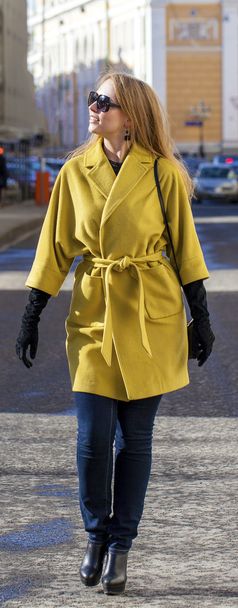 Young happy blonde woman in yellow coat walks on spring street - Photo, Image