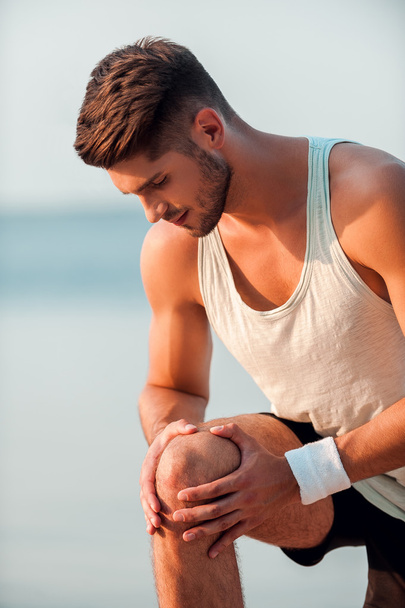 Feeling pain in his knee. Concentrated young muscular man massaging knee while standing outdoors - Fotoğraf, Görsel