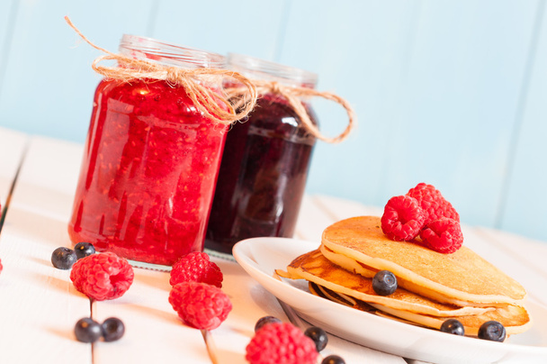 Stack of wheat golden pancakes or pancake cake with freshly picked raspberries on a dessert plate, glass mason jar full off blueberry and raspberry jam.  Unique perspective, selective focus - Fotó, kép