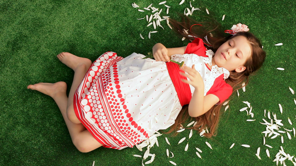 Девочка 6 лет лежит на траве и отделяется от ромашковых лепестков
. - Кадры, видео