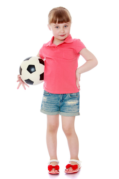 Adorable little blonde girl in denim shorts and red shirt is holding under his arm a soccer ball - Fotoğraf, Görsel