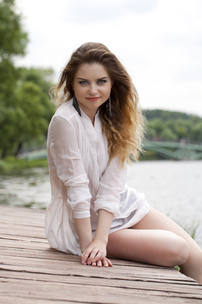 Young beautiful girl in a white tunic sits on a wooden pier on t - Photo, image