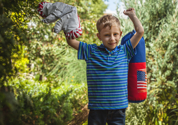Outdoor portrait of positive little boy in sunny summer garden with children's punching bag and gloves. - Foto, Imagem
