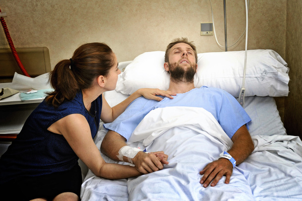 jovem casal no hospital quarto homem deitado na cama preocupado mulher segurando sua mão cuidando
 - Foto, Imagem