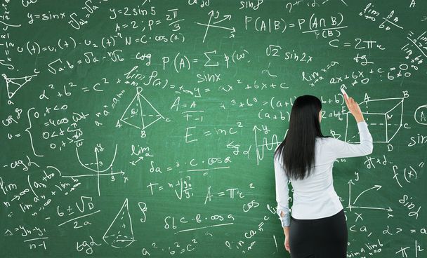 Rear view of a thoughtful woman who is writing math calculations on green chalk board. - Foto, Imagen