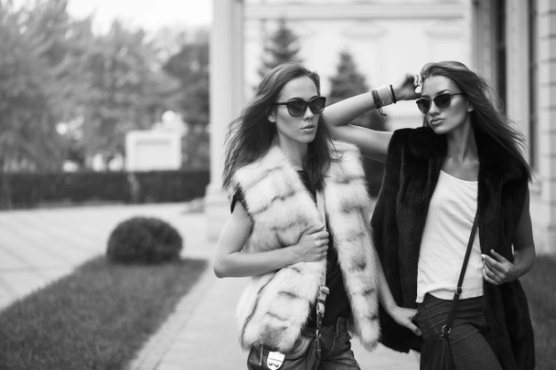 Fashion shot of two elegant beautiful girls in the sunset wearing sunglasses, fur vests. Две молодые женщины на улице. Вдохновение для покупок. Черно-белая концепция
 - Фото, изображение