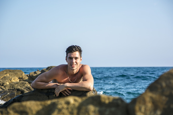 Young shirtless athletic man leaning on rock by sea - Фото, изображение