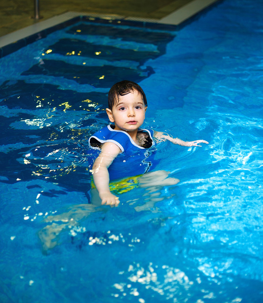 Мальчик с надувной плавательной жилеткой в бассейне, счастливая улыбка. Зрительный контакт
. - Фото, изображение