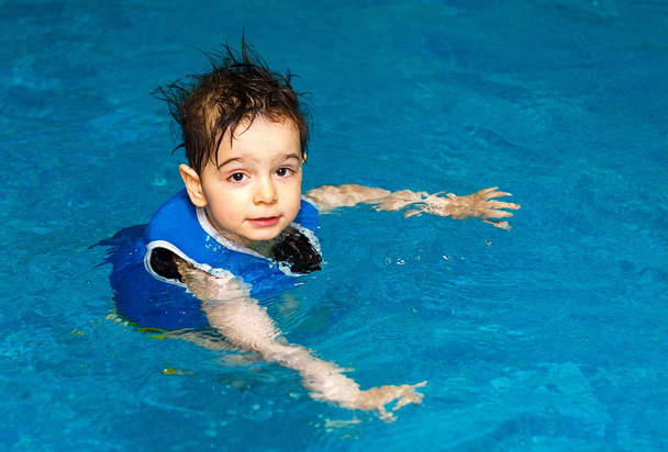 Niño con chaleco de natación inflable en la piscina, tiene una sonrisa feliz. Contacto visual
. - Foto, imagen