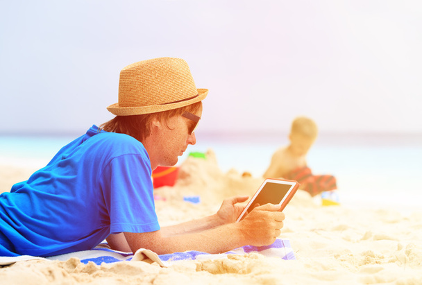 padre mirando touch pad mientras hijo jugar en la playa
 - Foto, imagen