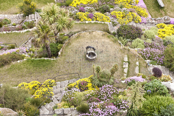 Blick von oben auf einen gepflegten bunten Garten mit Alpenblumen, Palmen, blühenden Sträuchern, verbrannt von Sonnengras und Zierwasser gut durch Steintreppe erreichbar - Foto, Bild
