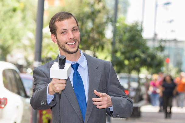 Attractive professional male news reporter wearing grey suit holding microphone, talking to camera from urban setting - Photo, Image