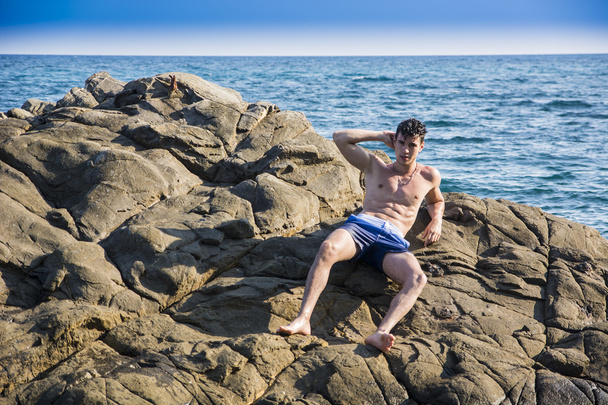 Young shirtless athletic man leaning on rock by water on ocean shore - Zdjęcie, obraz