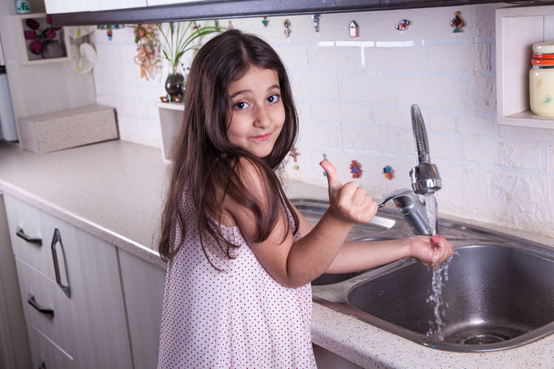 One beautiful middle eastern little girl with pink dress and long dark brown hair and eyes on white kitchen,helping parents to wash dishes and drinking water and smiling looking at camera. - Foto, imagen
