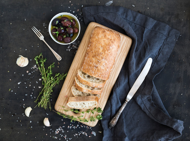 Italian ciabatta bread cut in slices on wooden chopping board with herbs, garlic and olives over dark grunge backdrop, copy space - Photo, Image