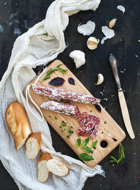 Meat gourmet snack. Salami, garlic, baguette and herbs on rustic wooden board over dark grunge backdrop - Photo, Image