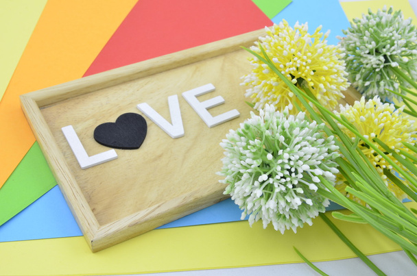 palabra blanca y símbolo de amor negro sobre fondo colorido.flor artificial verde, amarillo y blanco colocado a la derecha
 - Foto, Imagen