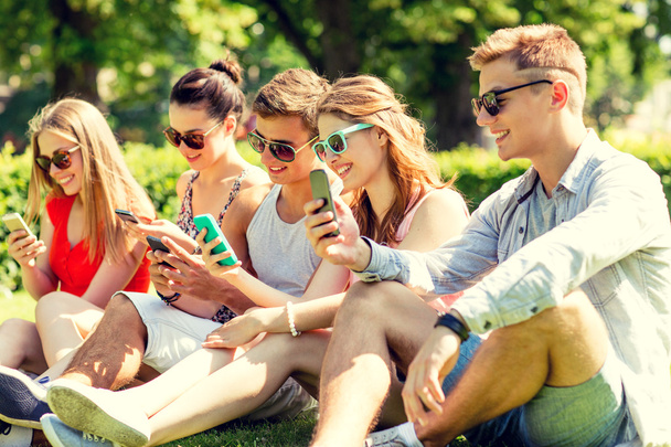 smiling friends with smartphones sitting on grass - Fotoğraf, Görsel