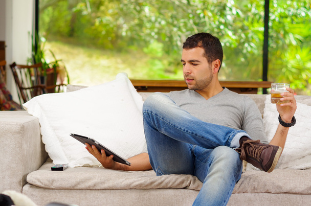 Hispanic man wearing denim jeans and grey sweater sitting on sofa laidback position holding a tablet with right hand, dark colored beverage glass in left - Zdjęcie, obraz