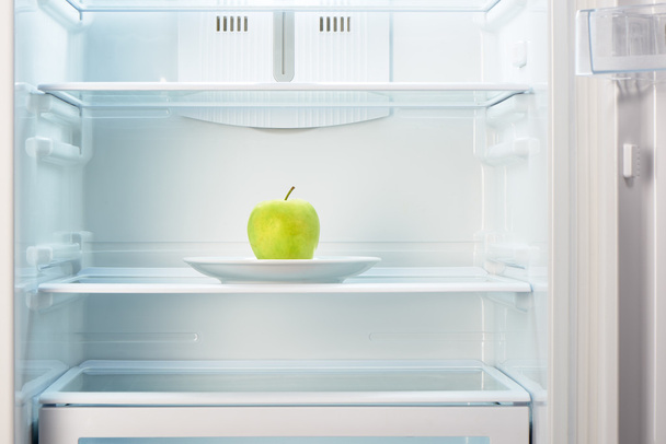 Green apple on white plate in open empty refrigerator - Foto, afbeelding