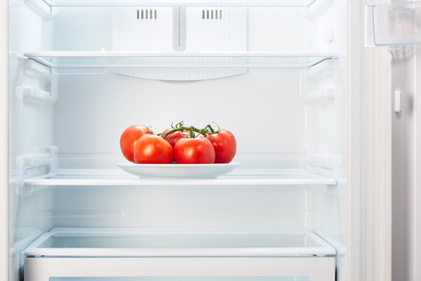 Branch of red tomatoes on white plate in open empty refrigerator - Foto, Imagen
