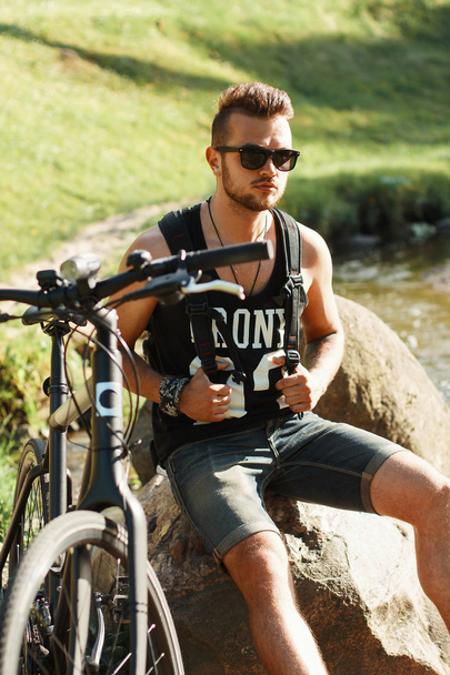 Kayanın üzerinde oturan bir bisiklet ile şık genç adam. Siyah T-shirt - Fotoğraf, Görsel