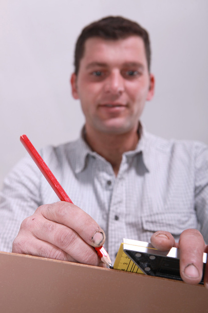 lähikuva miehestä, joka mittaa puunpalan ja merkitsee sen kynällä
 - Valokuva, kuva