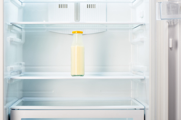 Glass bottle of yoghurt on shelf of open empty refrigerator - Фото, изображение