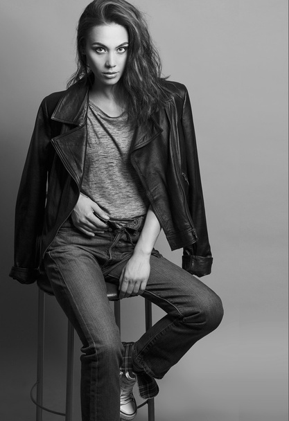 Portrait of a fashionable model with natural make up and perfect skin, dressed in men's jeans, grey shirt, black jacket and sneakers.  Studio shot. High fashion look. Monochrome (black and white)  photo - Fotó, kép