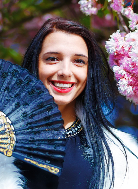 Hermosa mujer con flores, glamour de piel blanca y abanico negro en la mano, posando junto a flores mágicas de primavera rosa sakura. Fondo de flores
 - Foto, Imagen