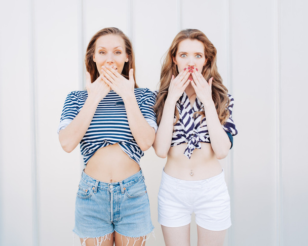 Portrait of two beautiful fashionable girlfriends in denim shorts and striped t-shirt posing nex to the glass wall. Have positive surprised emotions, screaming and laughing, bright summer day. - Photo, image