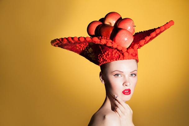 Creative beauty portrait of young beautiful woman looking at camera and posing in studio at big red hat with red apples. - Zdjęcie, obraz