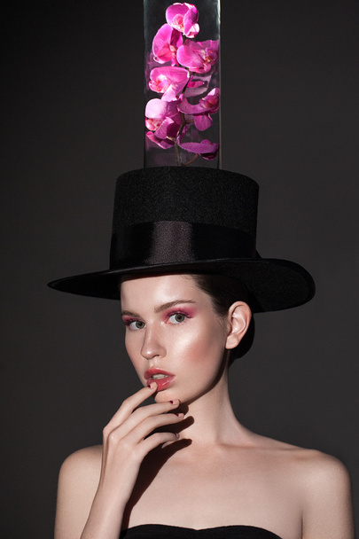 Portrait of attractive young woman looking at camera posing at studio in black hat with lilac pink orchid. - Fotoğraf, Görsel