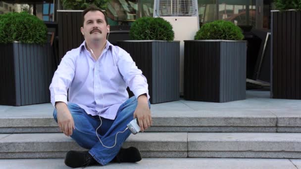 Geschäftsmann mittleren Alters hört Musik vom Smartphone sitzend auf den Stufen - Filmmaterial, Video