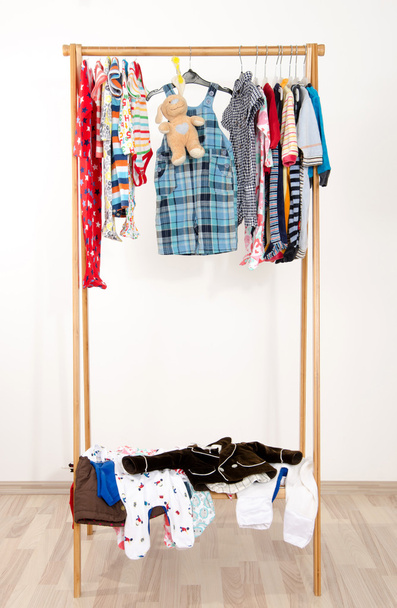 Dressing closet with clothes arranged on hangers.Colorful wardrobe of newborn,kids, toddlers, babies on a rack.Many t-shirts,pants, shirts,blouses, onesie hanging. Messy clothes thrown on a shelf - Фото, изображение