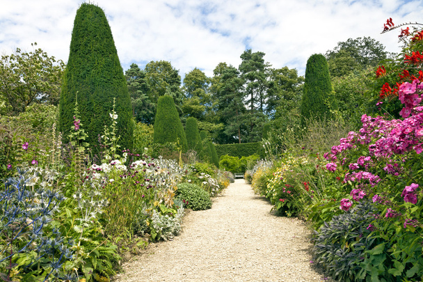 Stone pathway leading to a white bench, with cottage colourful flowers in bloom on both sides, shaped conifers, shrubs and tall trees in an English garden on a sunny summer day - Photo, Image
