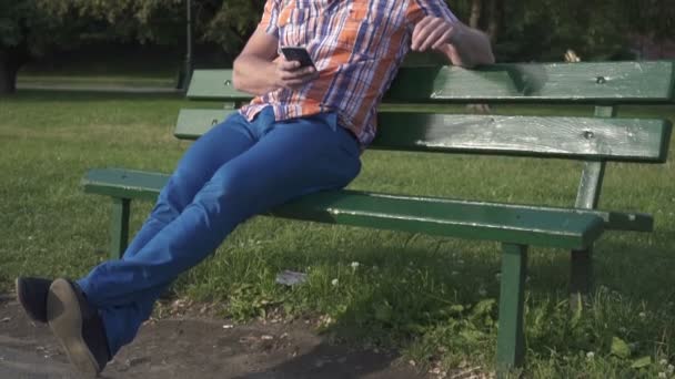 Mężczyzna przeglądający smartphone, siedząc na drewno brench, tilt i pan strzał - Materiał filmowy, wideo