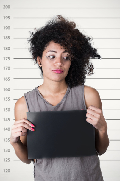 Hispanic brunette model with afro like hair wearing grey sleeveless shirt holding up blank board as posing for mugshot concept - Фото, изображение