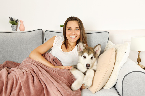 Young woman lying with malamute dog on sofa in room - Photo, Image