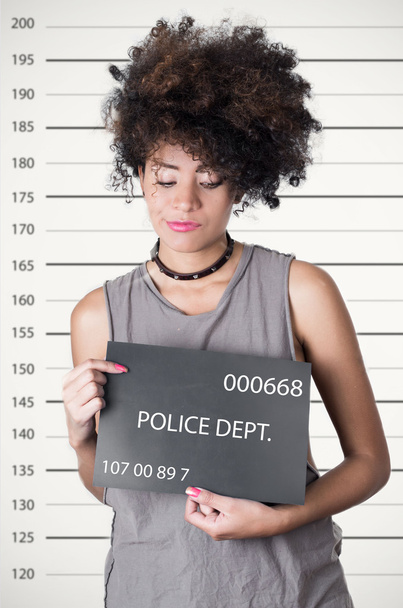 Hispanic brunette rebel model afro like hair wearing grey sleeveless shirt holding up police department board with number as posing for mugshot, careless facial expression - Foto, Bild