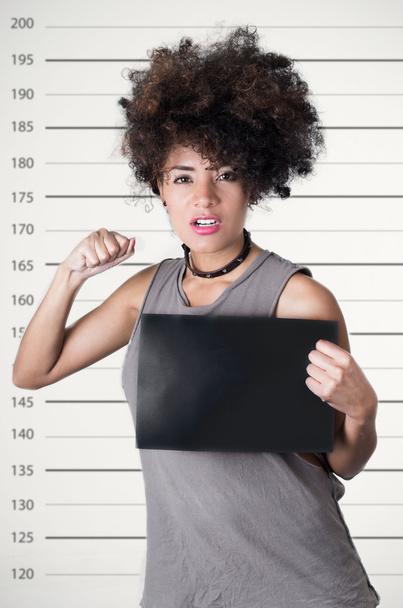 Hispanic brunette rebel model with afro like hair wearing grey sleeveless shirt holding up blank board as posing for mugshot concept, lifting one arm simulating punch - Foto, immagini